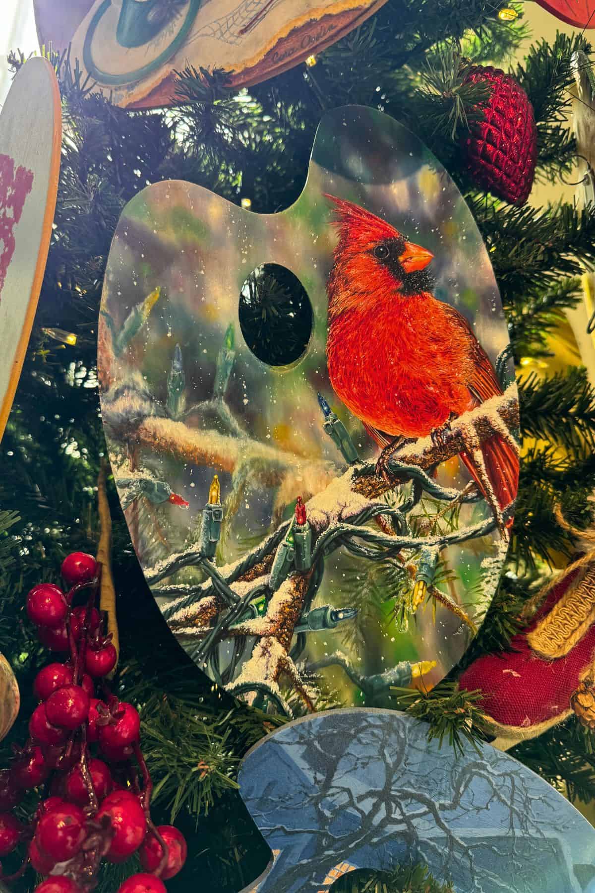 A painted artist's palette ornament featuring a cardinal on a snowy branch hangs on a decorated Christmas tree.