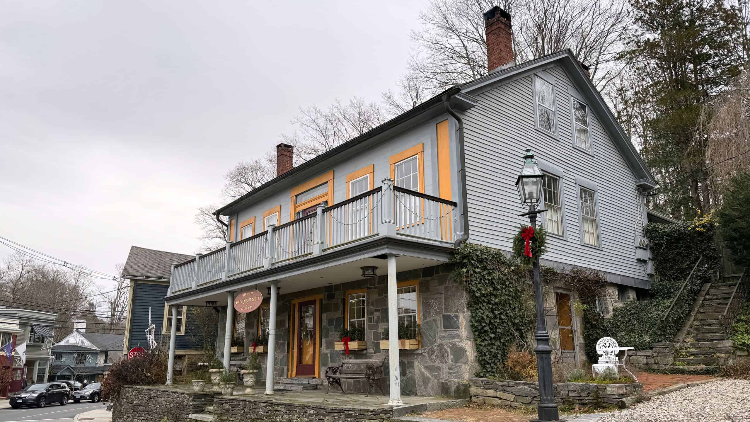 A two-story gray building with yellow trim, stone foundation, and a balcony, located on a street corner with bare trees and overcast sky.