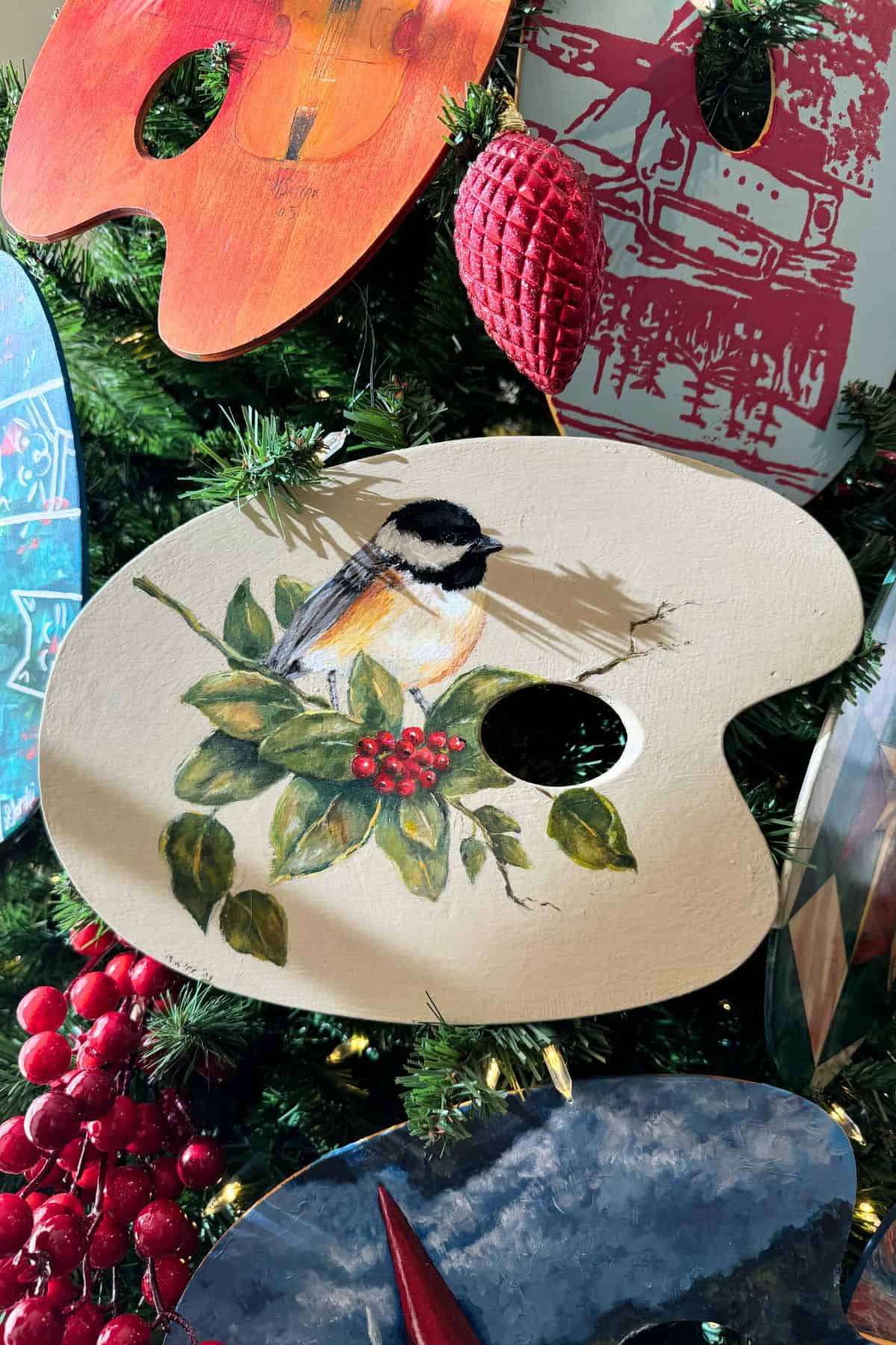 A painted artist palette on a Christmas tree features a bird perched on a branch with red berries, surrounded by other decorated palettes and festive ornaments.