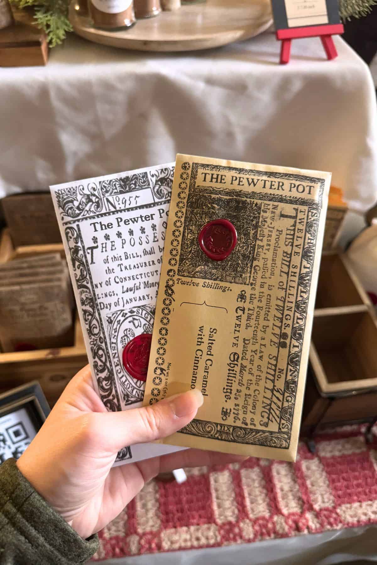 A hand holds two vintage-style packages labeled "The Pewter Pot," featuring decorative text and red wax seals. A patterned rug and wooden boxes are in the background.