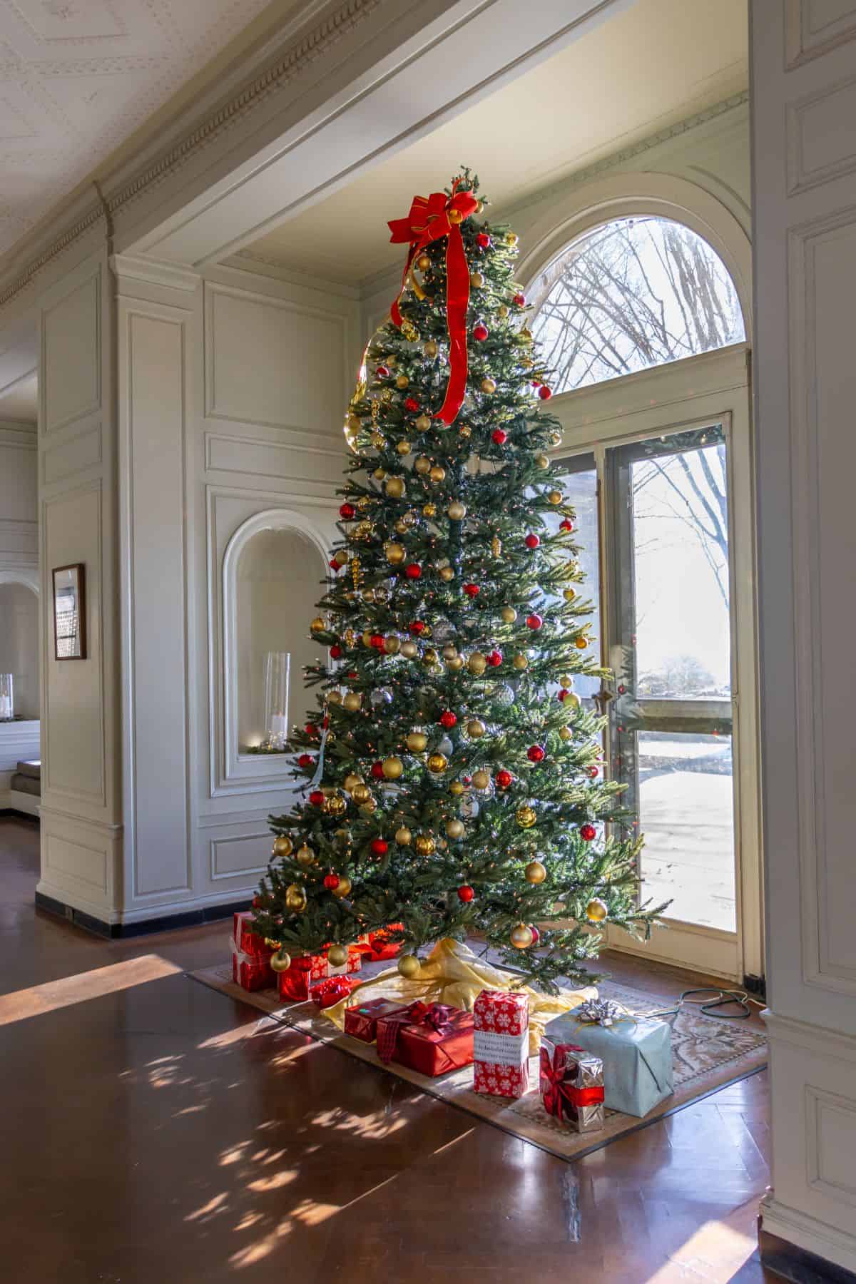 A decorated Christmas tree with a red bow on top stands by a window, surrounded by wrapped presents on the floor in a bright, elegant room.