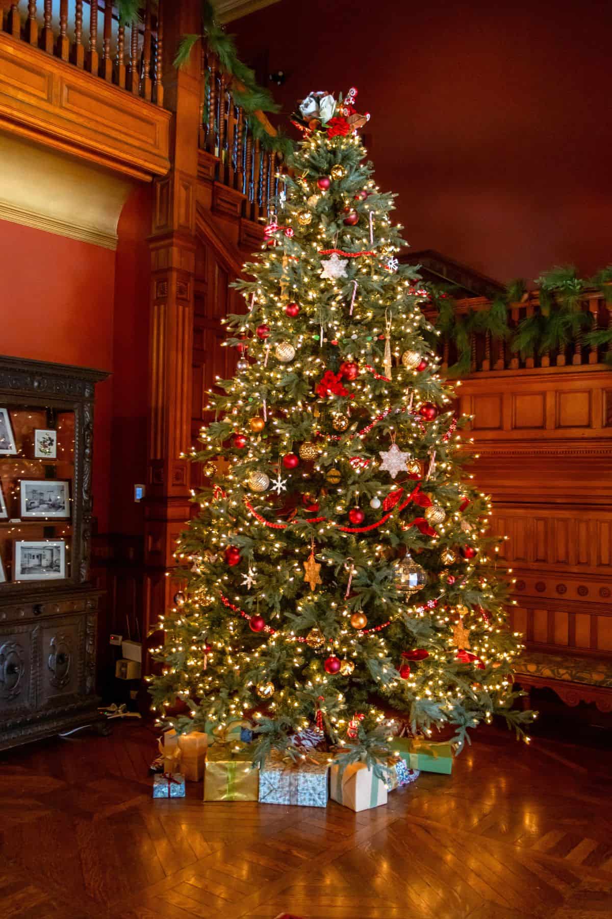 A decorated Christmas tree with lights, ornaments, and wrapped gifts underneath, standing in a wood-paneled room with garland on the banister.