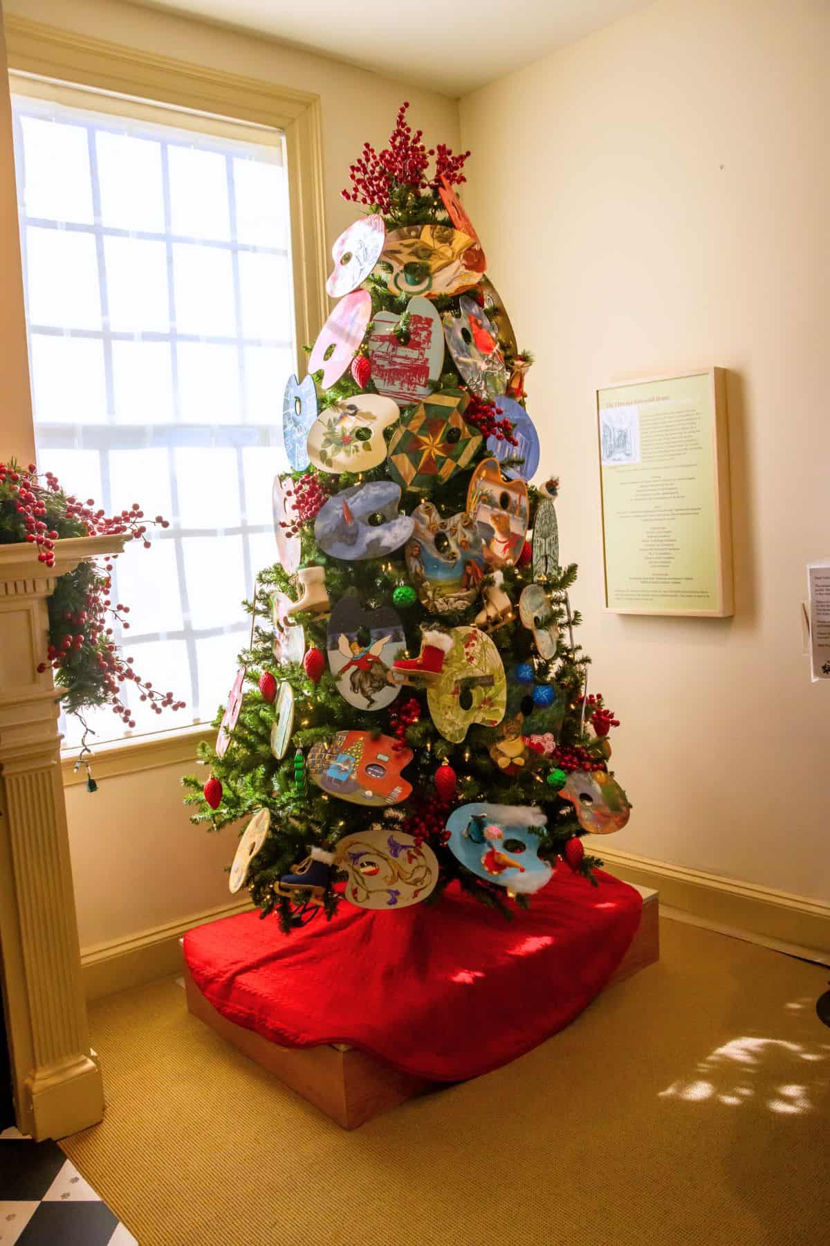 Artificial Christmas tree decorated with handmade paper ornaments, red berries, and green baubles, placed on a red tree skirt near a window in a well-lit room.