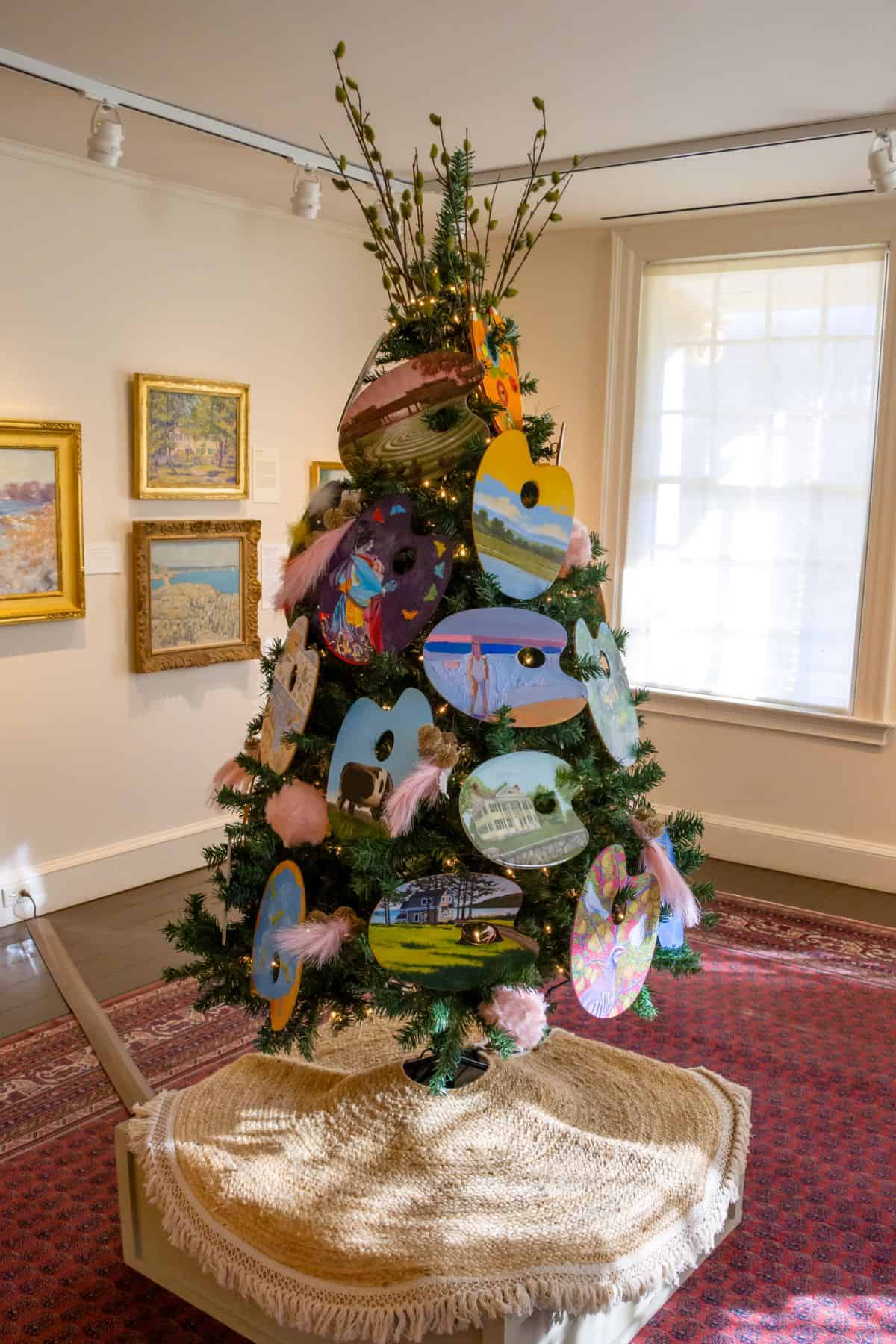 Decorated Christmas tree in an art gallery, adorned with artist palette ornaments featuring painted scenes, surrounded by framed paintings on the walls.