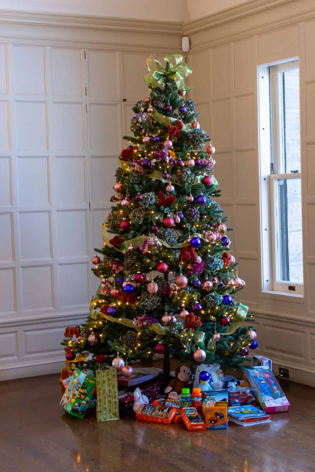 A decorated Christmas tree with ornaments and lights stands in a corner near a window, with various wrapped and unwrapped toys and gifts placed underneath.
