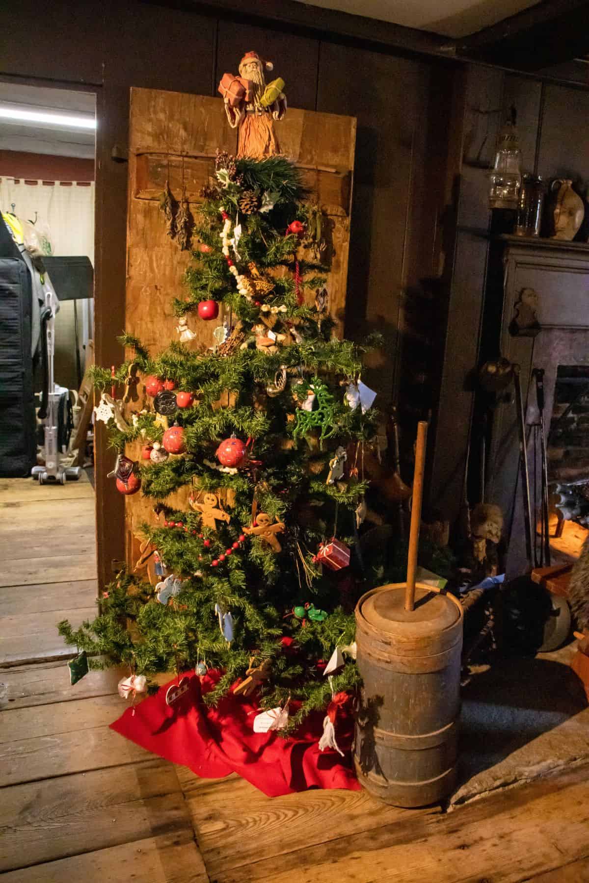 A decorated Christmas tree with a red fabric base stands beside an old wooden butter churn in a rustic room with wooden floors and a fireplace.