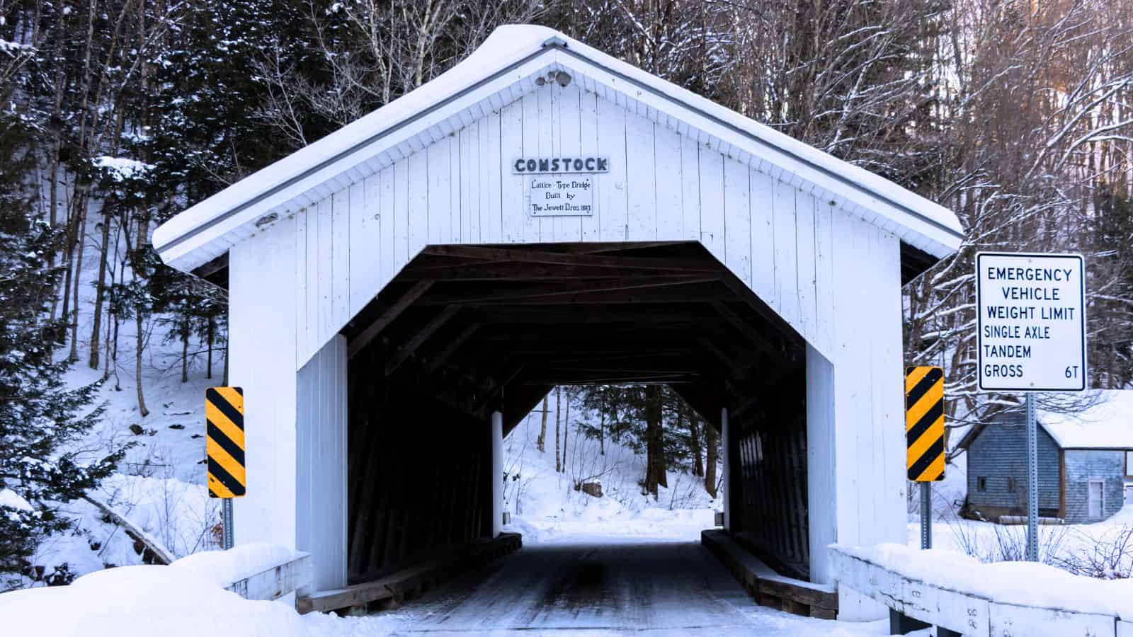 A white New England covered bridge stands in a snowy landscape, marked by caution signs at the entrance and a road sign indicating weight limits for emergency vehicles.