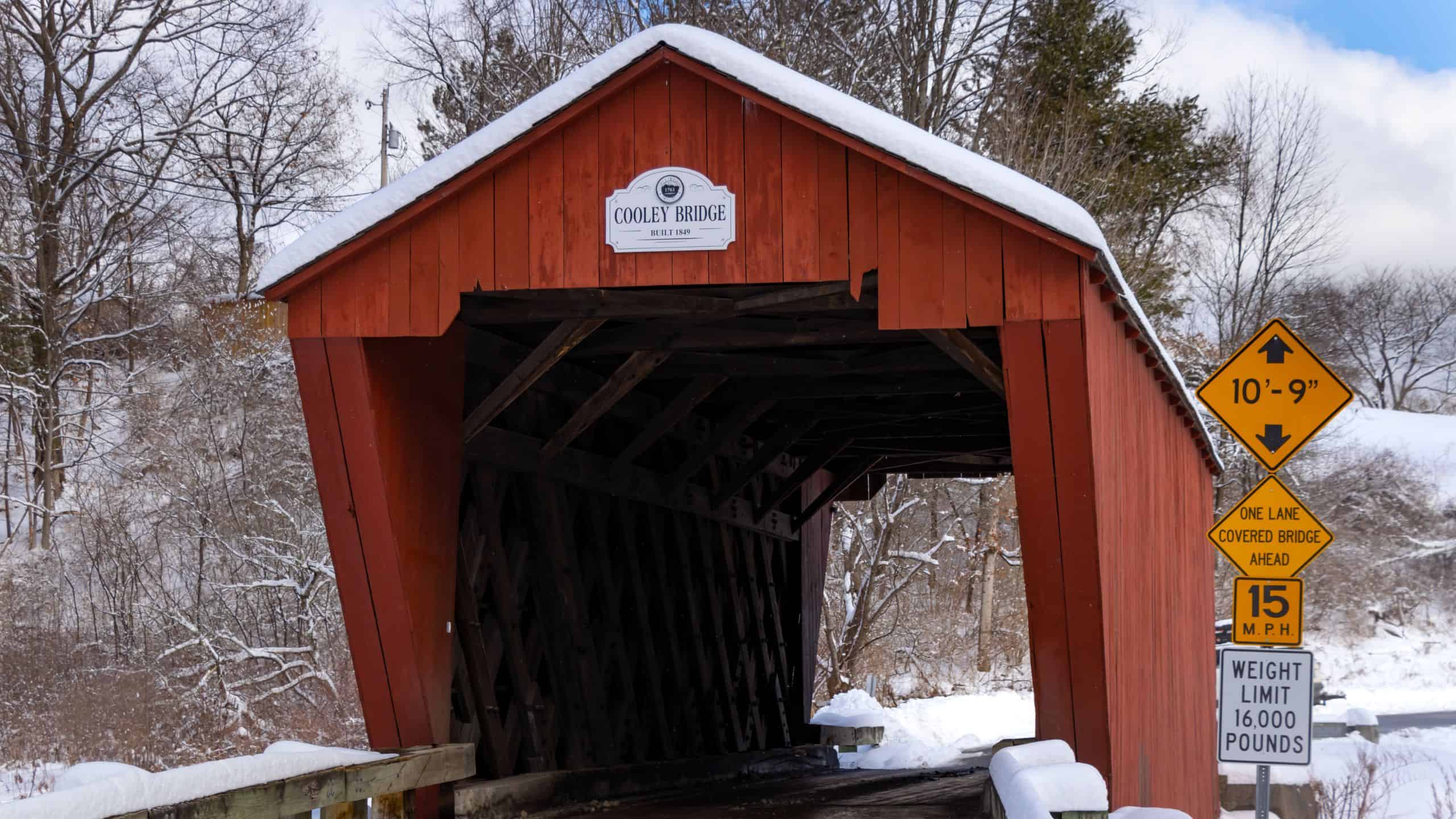 Red covered wooden bridge in snowy landscape with height restriction and weight limit traffic signs at entrance. Bridge sign reads "Coxey Bridge.