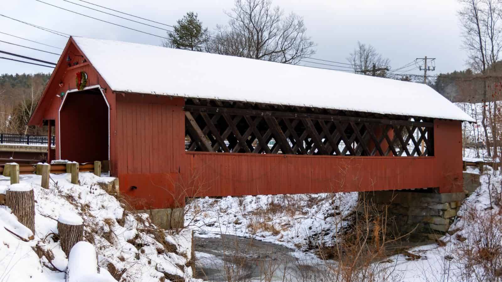 A red wooden New England covered bridge with a snow-covered roof spans a small stream in a serene winter landscape.