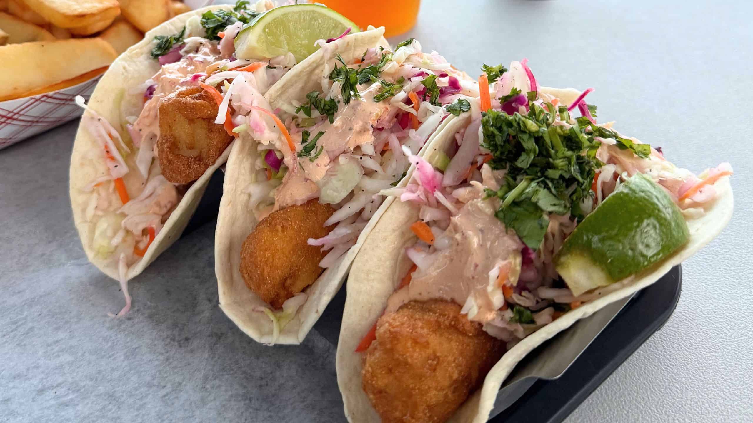 Three fish tacos with shredded cabbage, sauce, lime wedges, and chopped herbs in soft tortillas, accompanied by fries in the background.