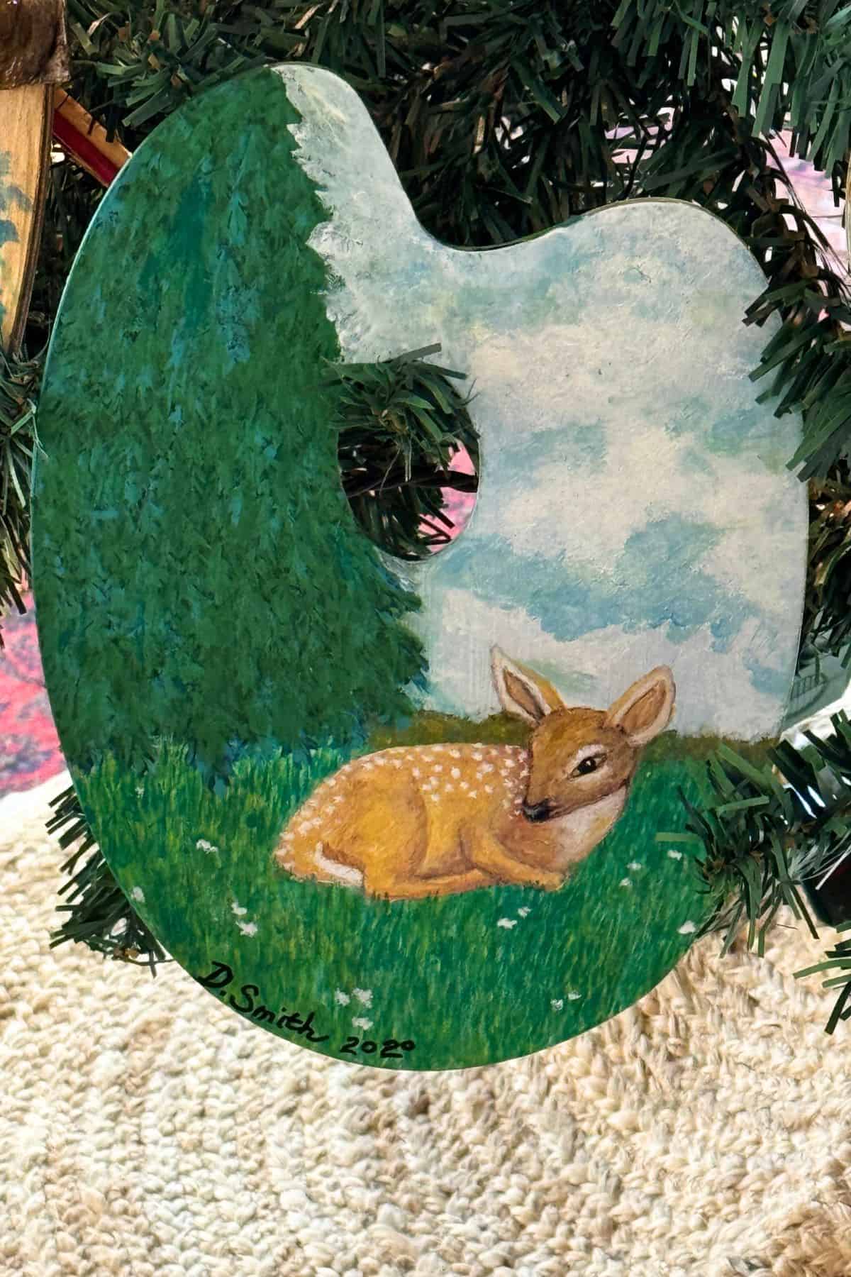 A painted ornament shaped like an artist’s palette hangs on a Christmas tree, featuring a fawn lying in grass beside trees and a cloudy sky. The ornament is signed “D. Smith 2022.”.