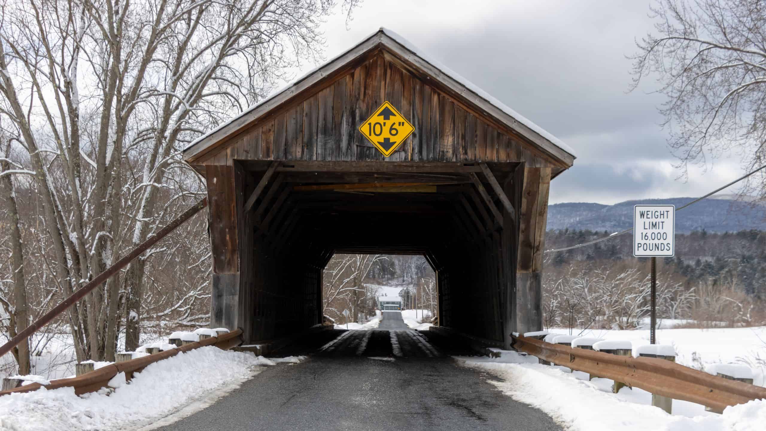 A wooden covered bridge in a snowy rural area with a height clearance sign of 10 feet 6 inches and a weight limit sign of 16,000 pounds.