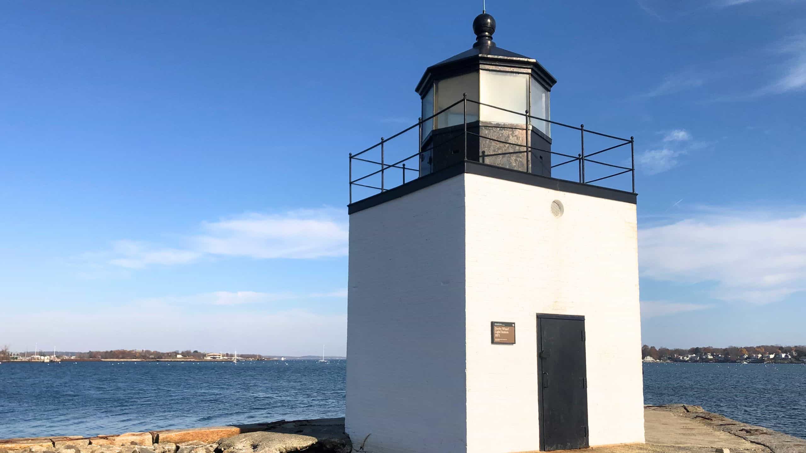 A square white lighthouse with a black lantern stands by the water, with a partly cloudy sky and shoreline visible in the background.