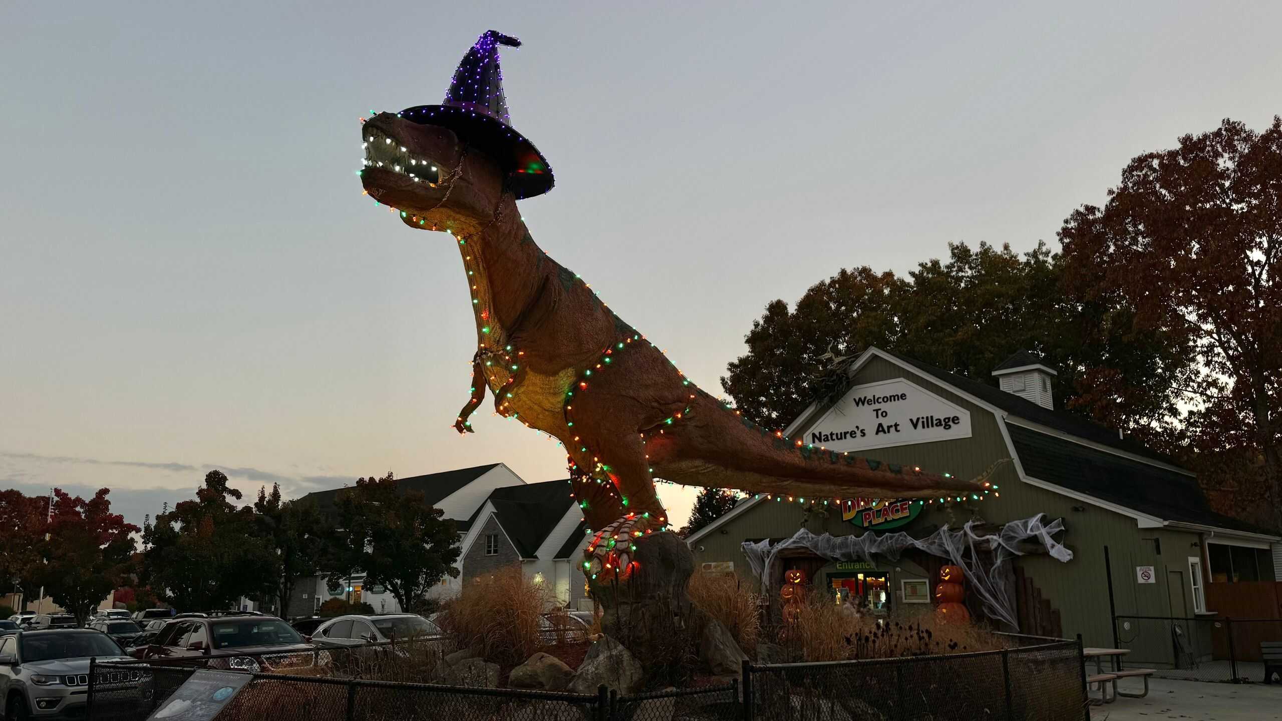A large dinosaur statue decorated with string lights and wearing a purple witch hat stands outside a building labeled "Nature's Art Village.