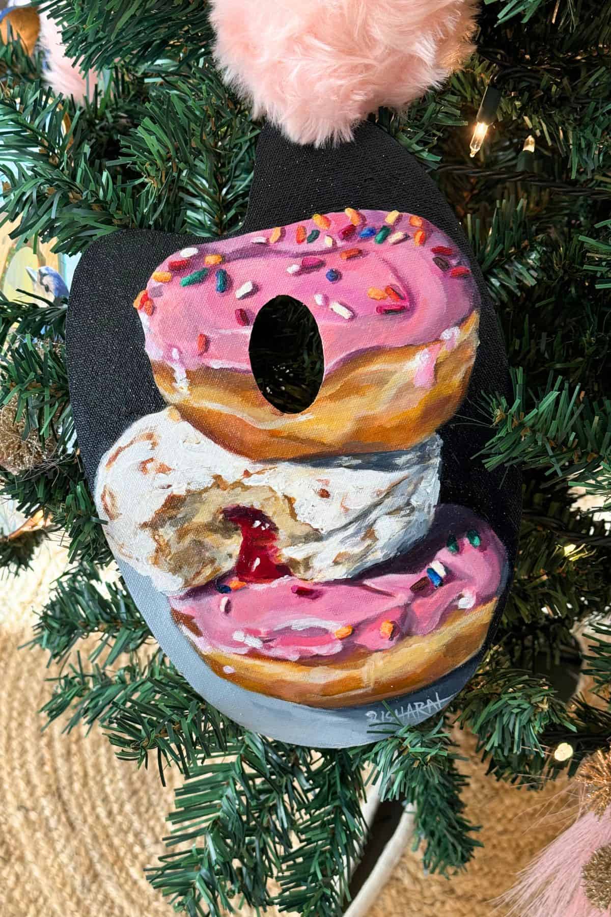 A painted ornament featuring three stacked donuts—two with pink icing and sprinkles, one with powdered sugar and jelly filling—hangs on a decorated Christmas tree.