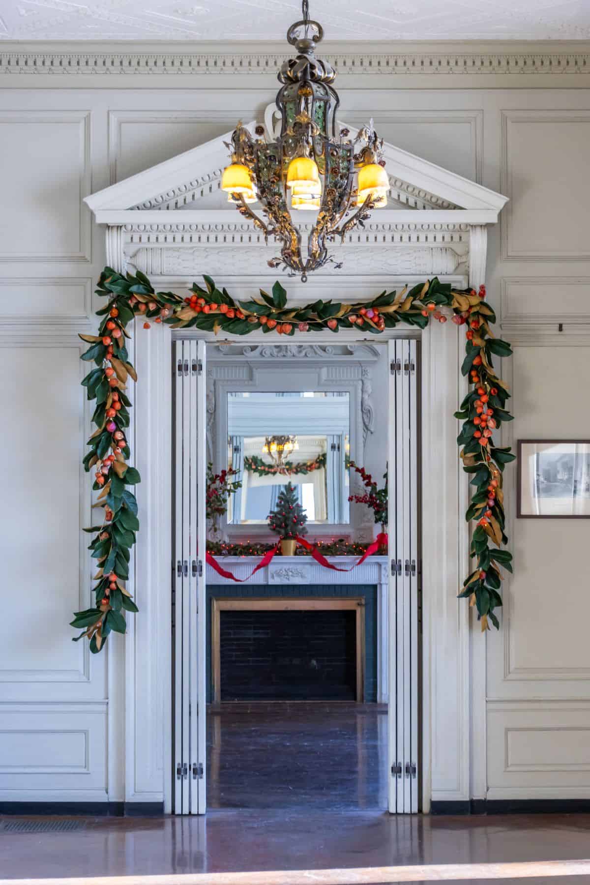 A chandelier hangs above a doorway decorated with garland, leading to a series of mirrored doorways and a fireplace with red holiday decorations.
