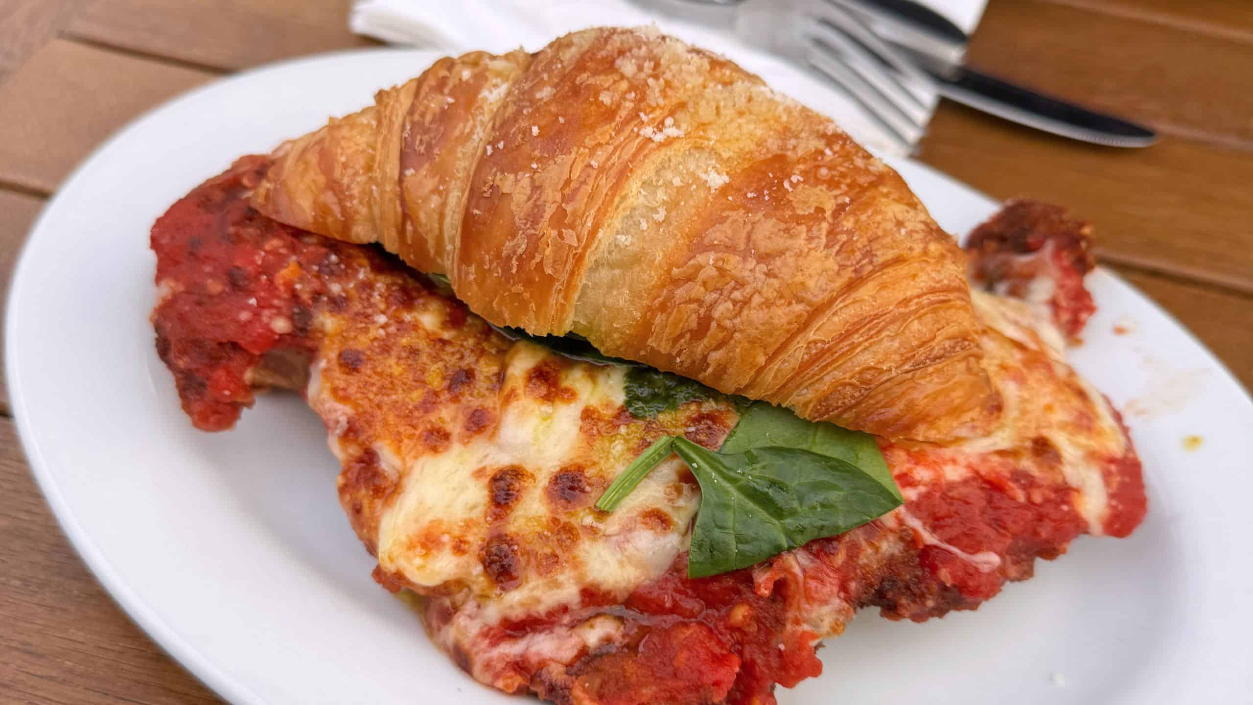 A croissant placed on top of a breaded, cheese-covered chicken parmesan with tomato sauce and spinach on a white plate.