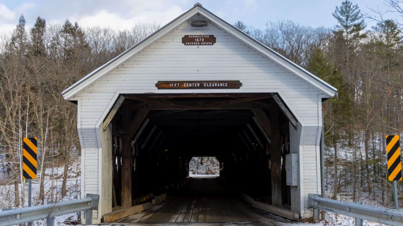 A white wooden covered bridge with a peaked roof stands over a snowy road, surrounded by leafless trees—a classic example of New England covered bridges. Caution and clearance signs are posted at the entrance.