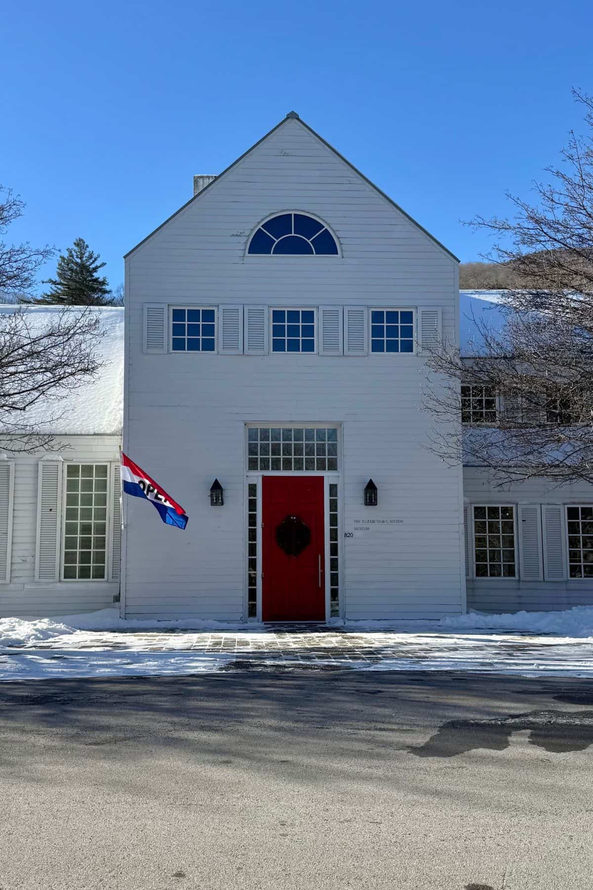 A white building with a red door, black wreath, and a flag that reads "OPEN" on a snowy ground under a clear blue sky.