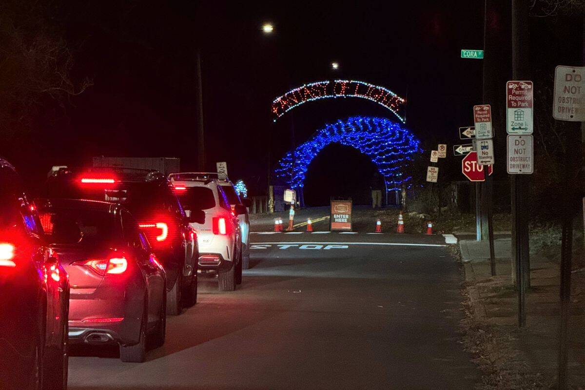 Cars wait in line at night to see a drive-through holiday light display with blue and red illuminated arches ahead.