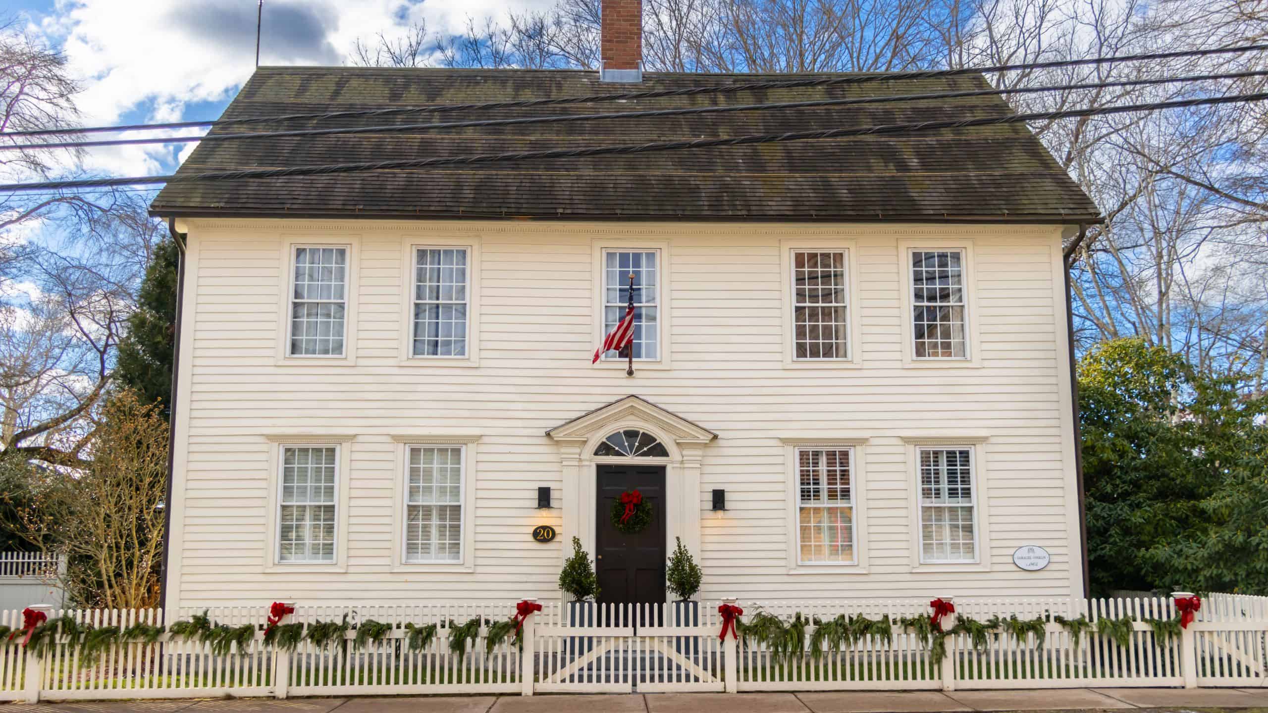 A two-story white colonial house with nine windows, a black door, a picket fence decorated with garlands and red bows, and an American flag above the entrance.