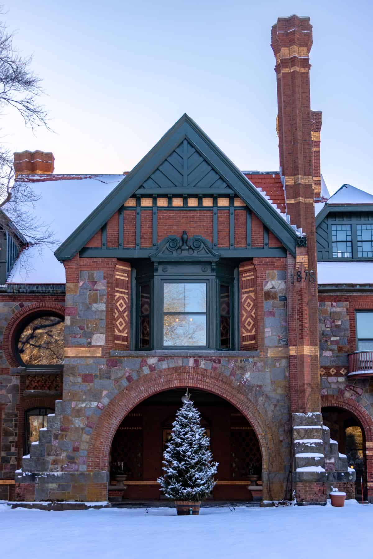 A stone and brick house with arched entryway, two tall chimneys, decorative brickwork, and a snow-covered evergreen tree in front. Snow is on the ground and roof.