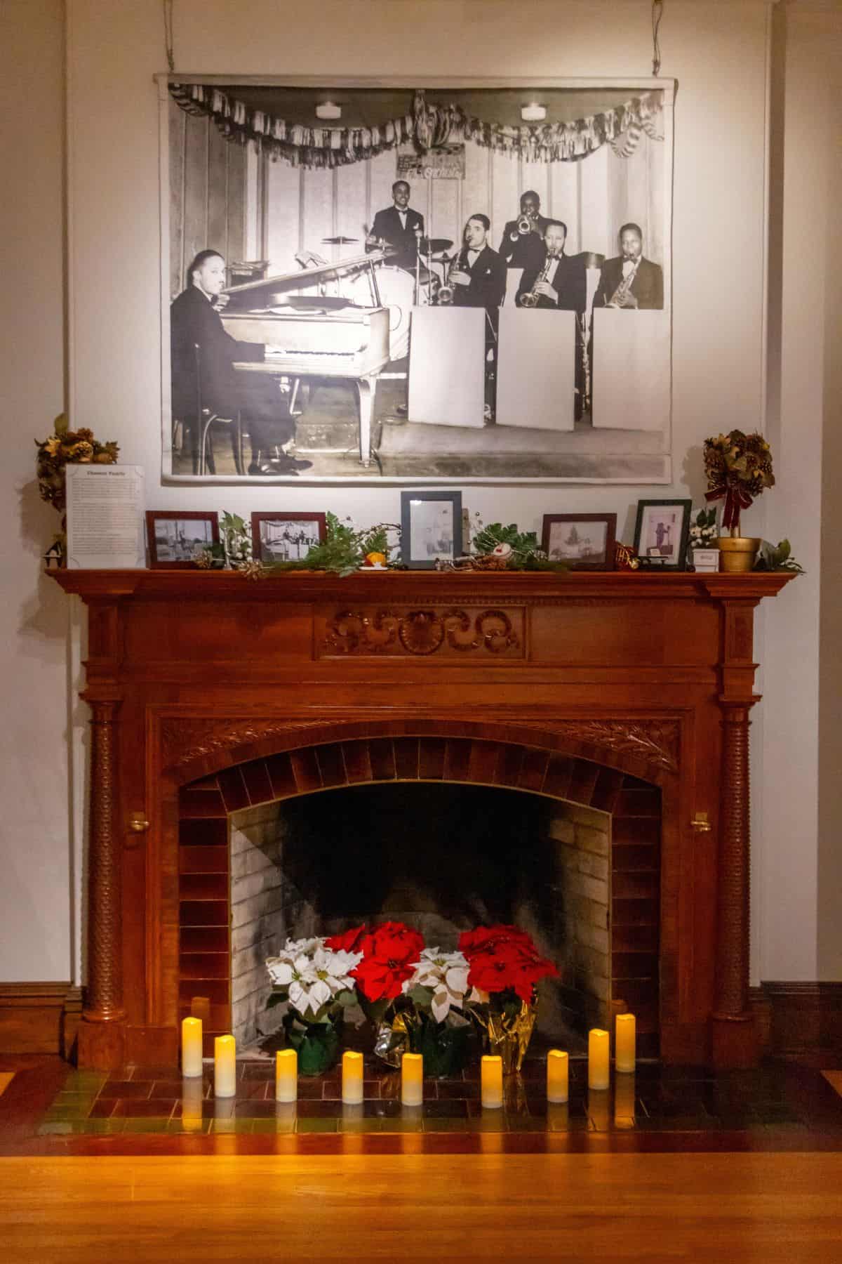 A wooden fireplace decorated with candles, poinsettias, framed photos, and garlands, with a large black-and-white photo of a jazz band hanging above.