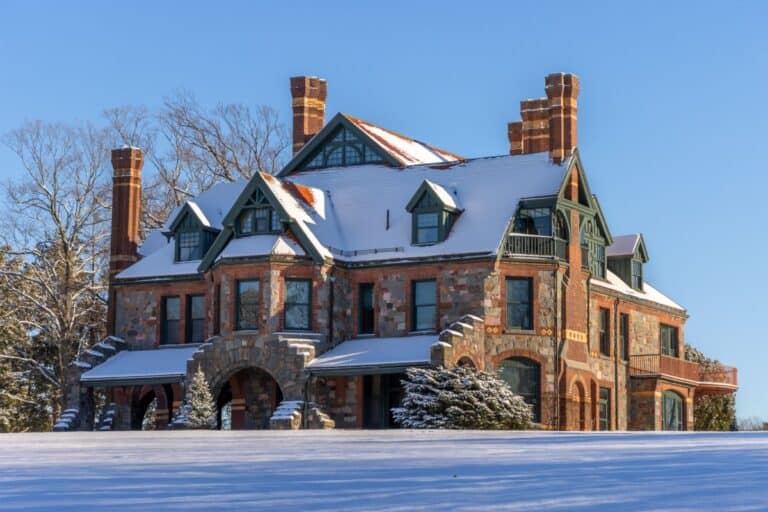 Large stone mansion with multiple chimneys and peaked roofs, covered in snow, set against a clear blue sky and surrounded by snow-covered ground.