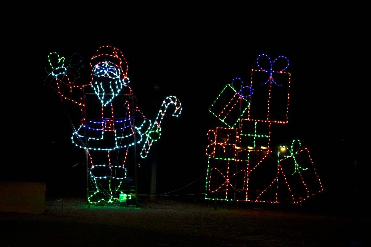 Neon light display of Santa Claus holding a candy cane and waving, next to a stack of colorful, gift-wrapped presents, set against a dark background.