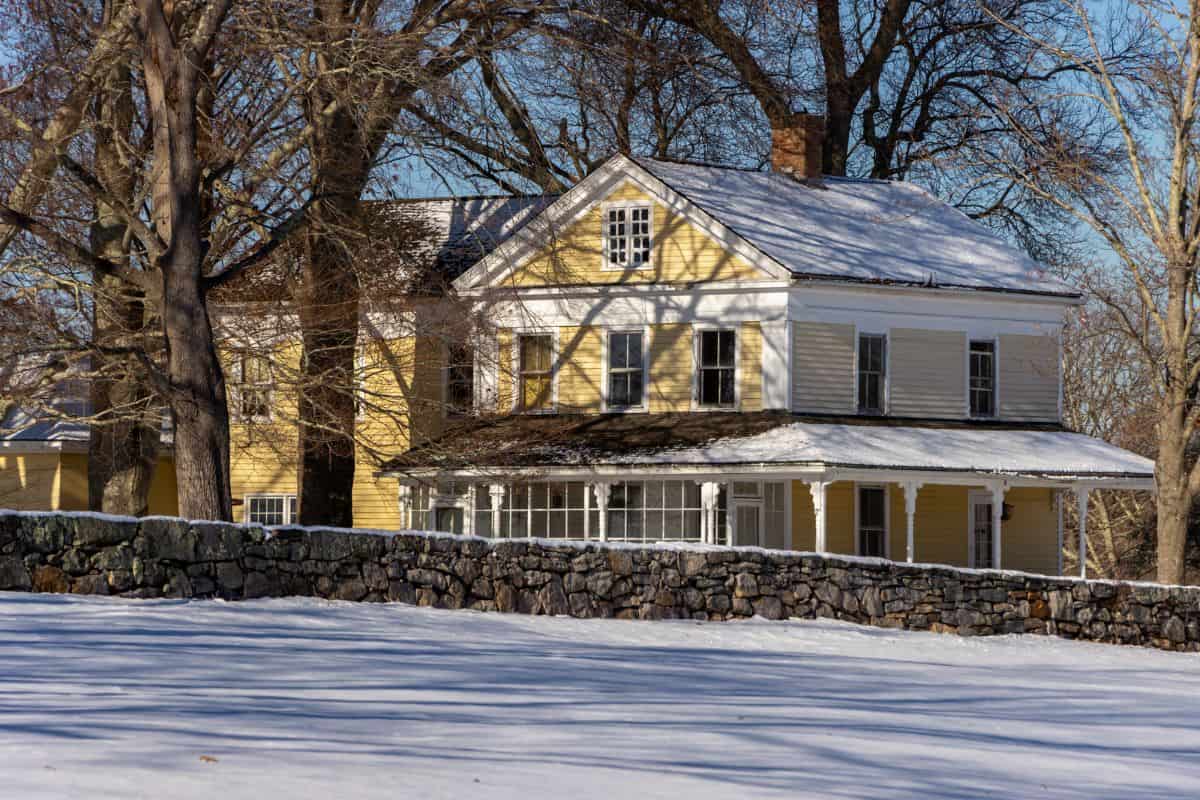 A large yellow house with a covered porch stands behind a stone wall, surrounded by leafless trees and snow-covered ground on a sunny winter day.