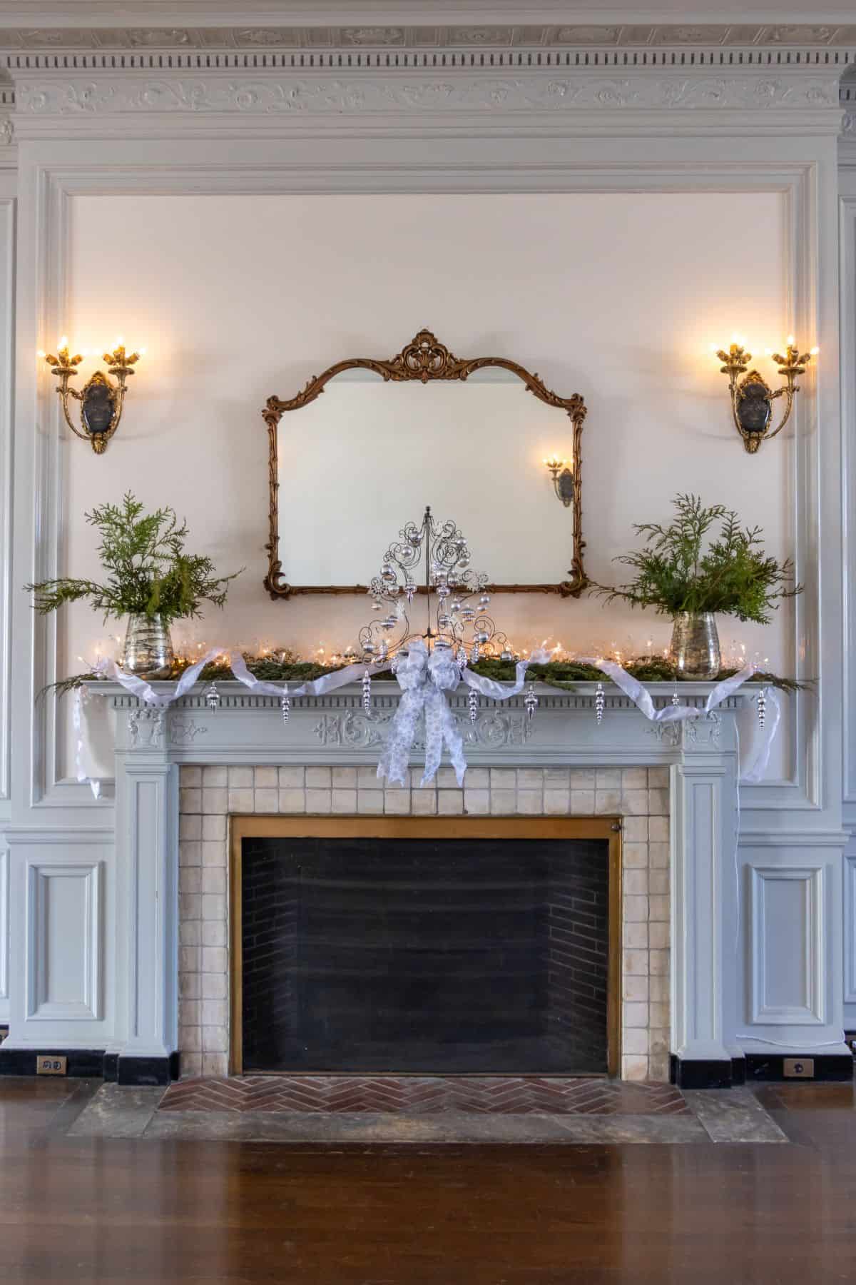 Ornate fireplace with a tiled hearth, decorated with greenery, white ribbon, string lights, and flanked by two wall sconces beneath a large mirror.