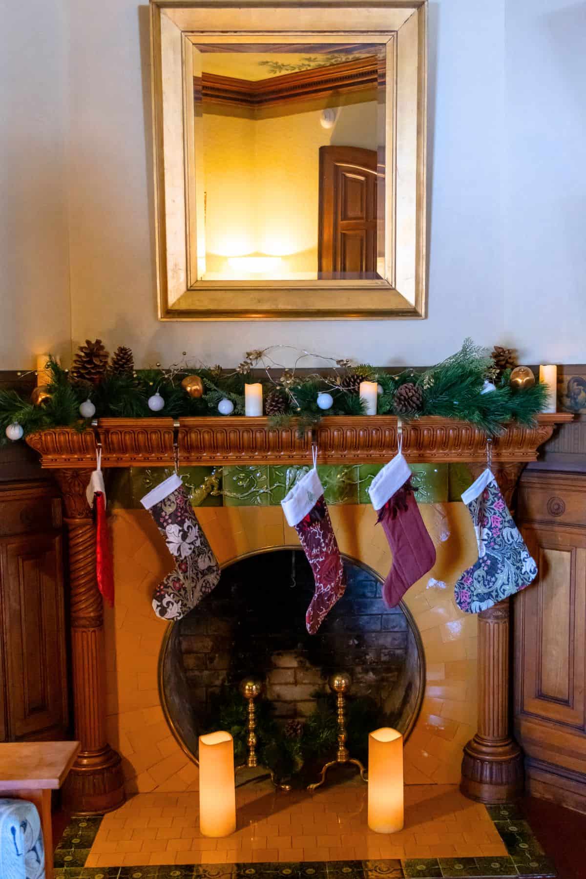 A decorated fireplace with four Christmas stockings, greenery, pinecones, candles, and a large gold-framed mirror above the mantel.