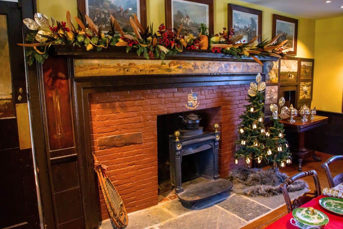 A brick fireplace with a decorated mantle, a small Christmas tree with lights, vintage snowshoes, and framed paintings on the wall in a cozy room.