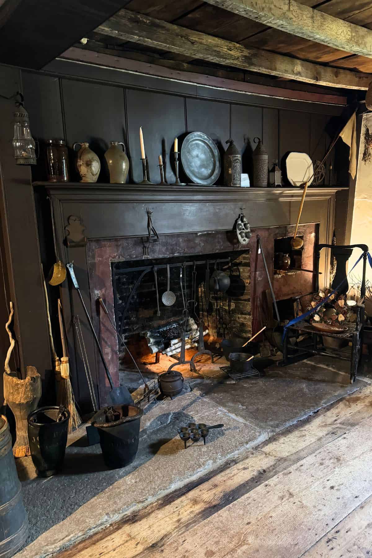 A large stone fireplace with cooking utensils, iron pots, buckets, and various ceramic jugs and metalware displayed on the mantel in a rustic, historic room.