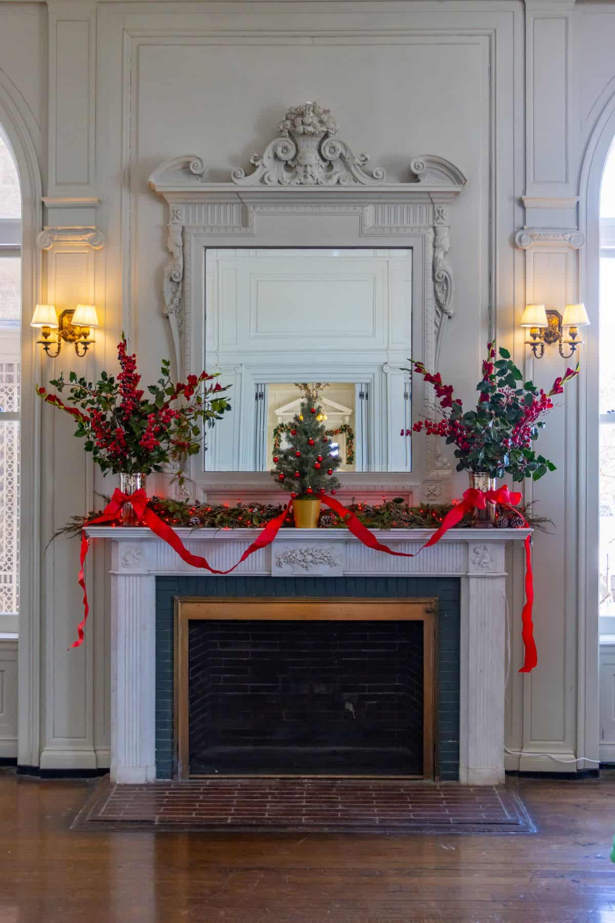 A white fireplace decorated with red ribbons, greenery, and red berries, with a small Christmas tree centerpiece and a large ornate mirror above the mantel.
