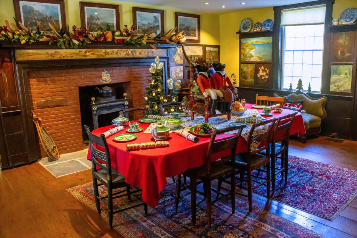 A dining room decorated for Christmas with a red tablecloth, festive ornaments, a small tree on the table, and garland above a brick fireplace. Paintings and plates decorate the walls.