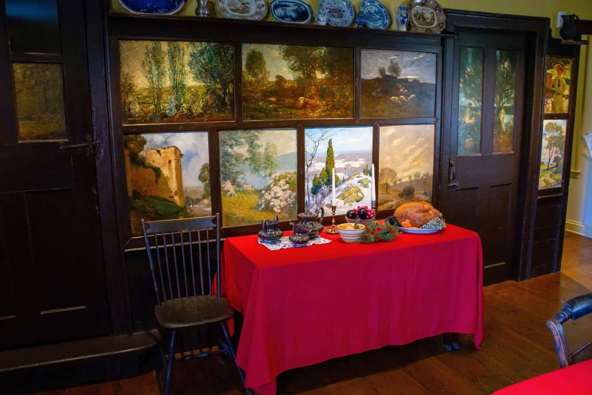 A table with a red tablecloth displays dishes, glassware, and a cooked ham in a room with dark wooden walls and landscape paintings.