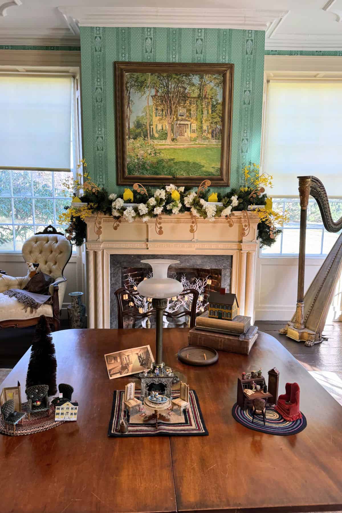 A decorated living room with a wooden table displaying miniature models, a white fireplace with flowers, a large painting above it, and a harp in the corner.