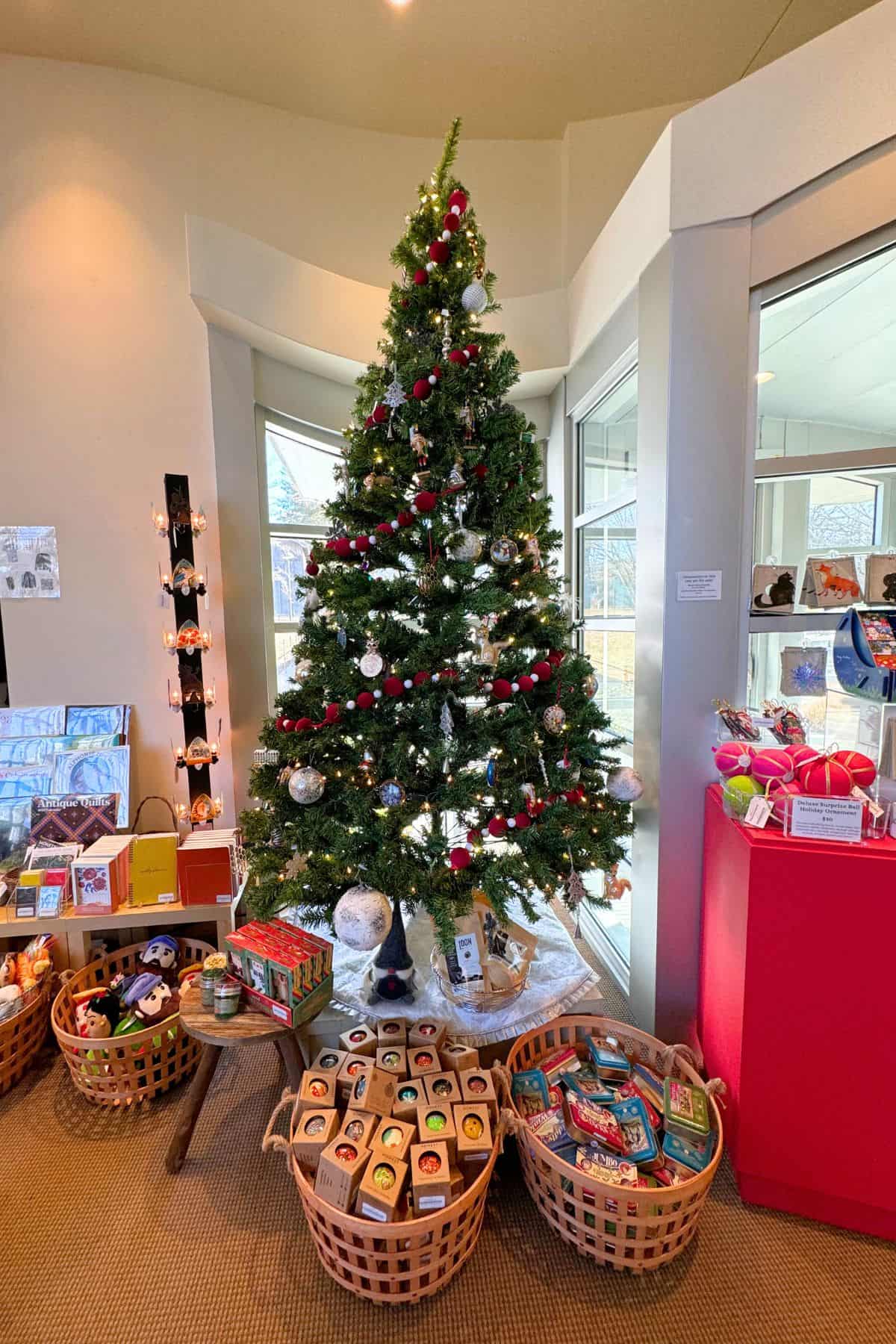 A decorated Christmas tree stands indoors, surrounded by baskets of packaged goods and toys, near a window and display shelves.