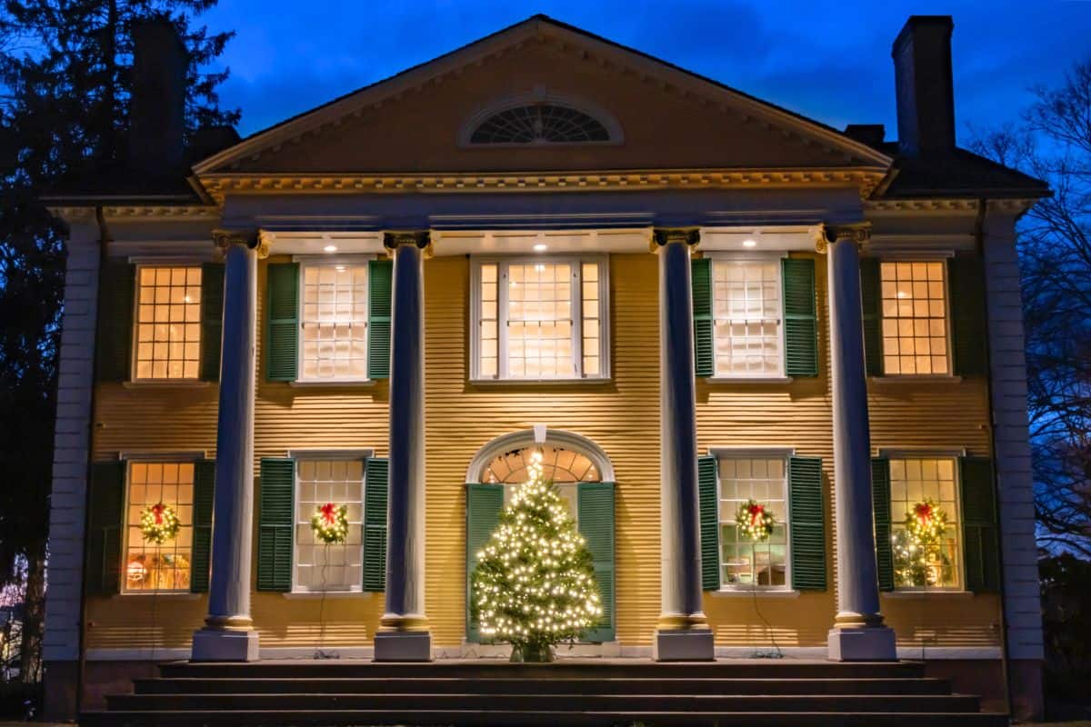 A yellow two-story house with white columns is decorated for Christmas with wreaths on the windows and a lit Christmas tree in front, photographed at dusk.