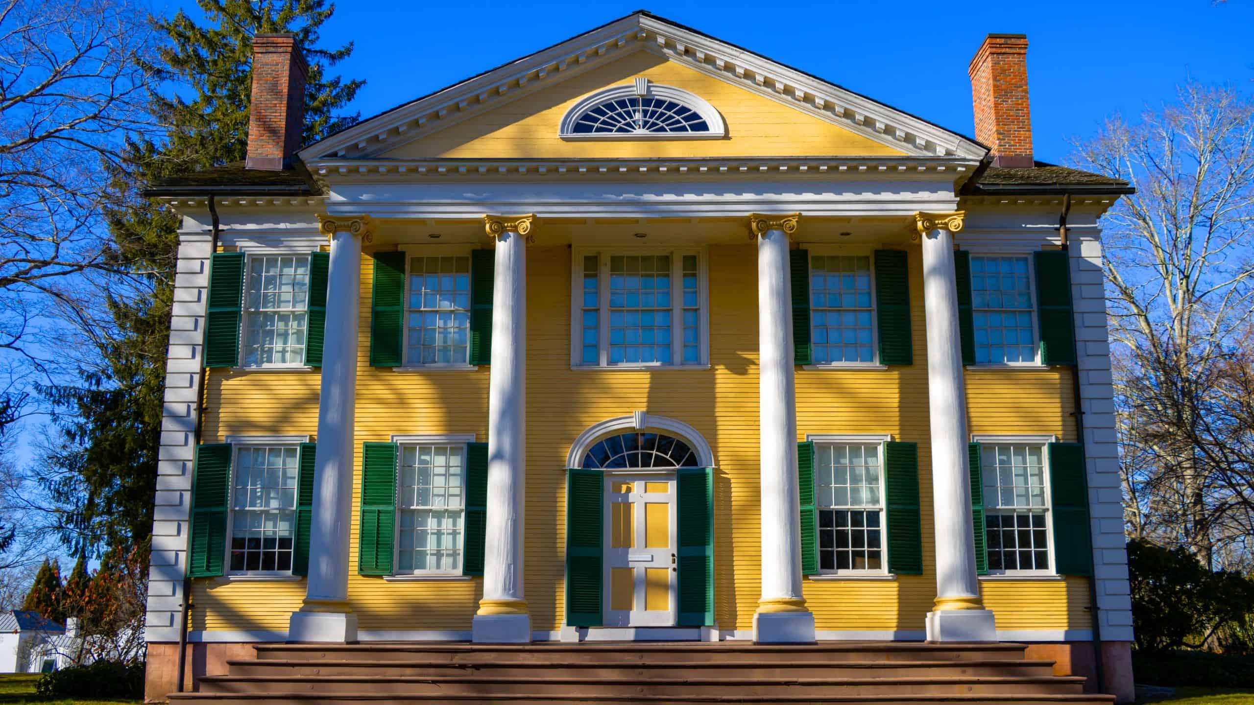 A yellow two-story house with white columns, green shutters, and a triangular pediment, set against a clear blue sky.