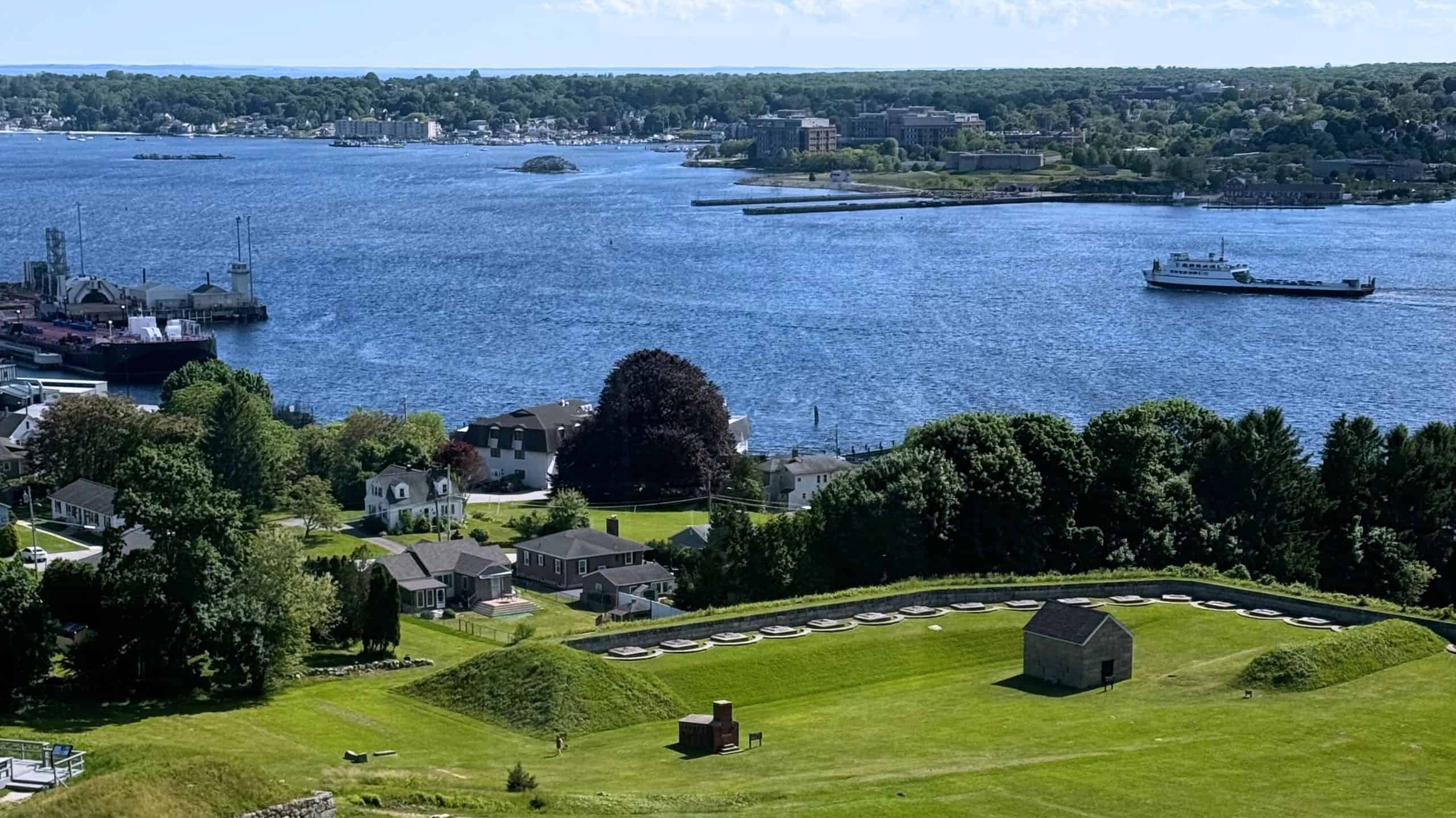 A view of a river with a ferry, surrounded by houses, green lawns, and trees; historical structures are visible in the foreground.