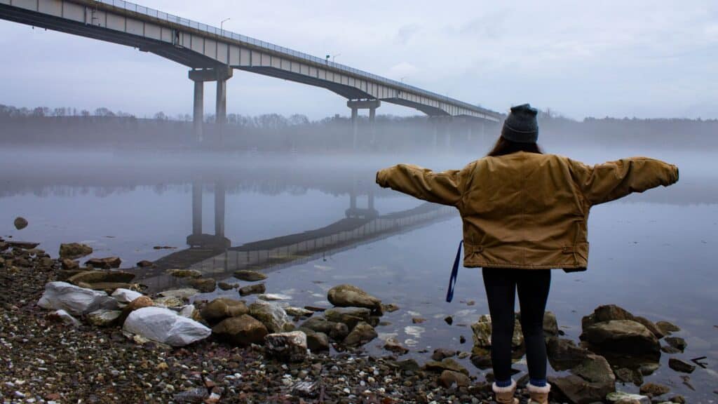 Person in a brown jacket stands on a rocky shore with arms outstretched, facing a foggy river and a bridge extending across the water.