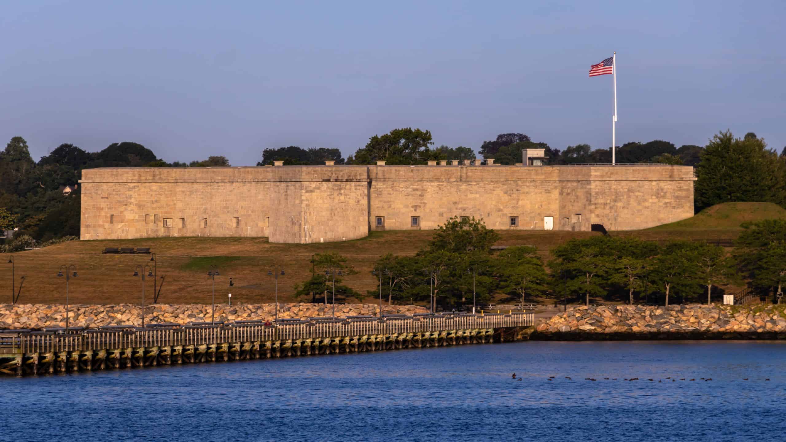 Rectangular stone fort with an American flag on a pole, surrounded by trees and water in the foreground, under a clear sky.