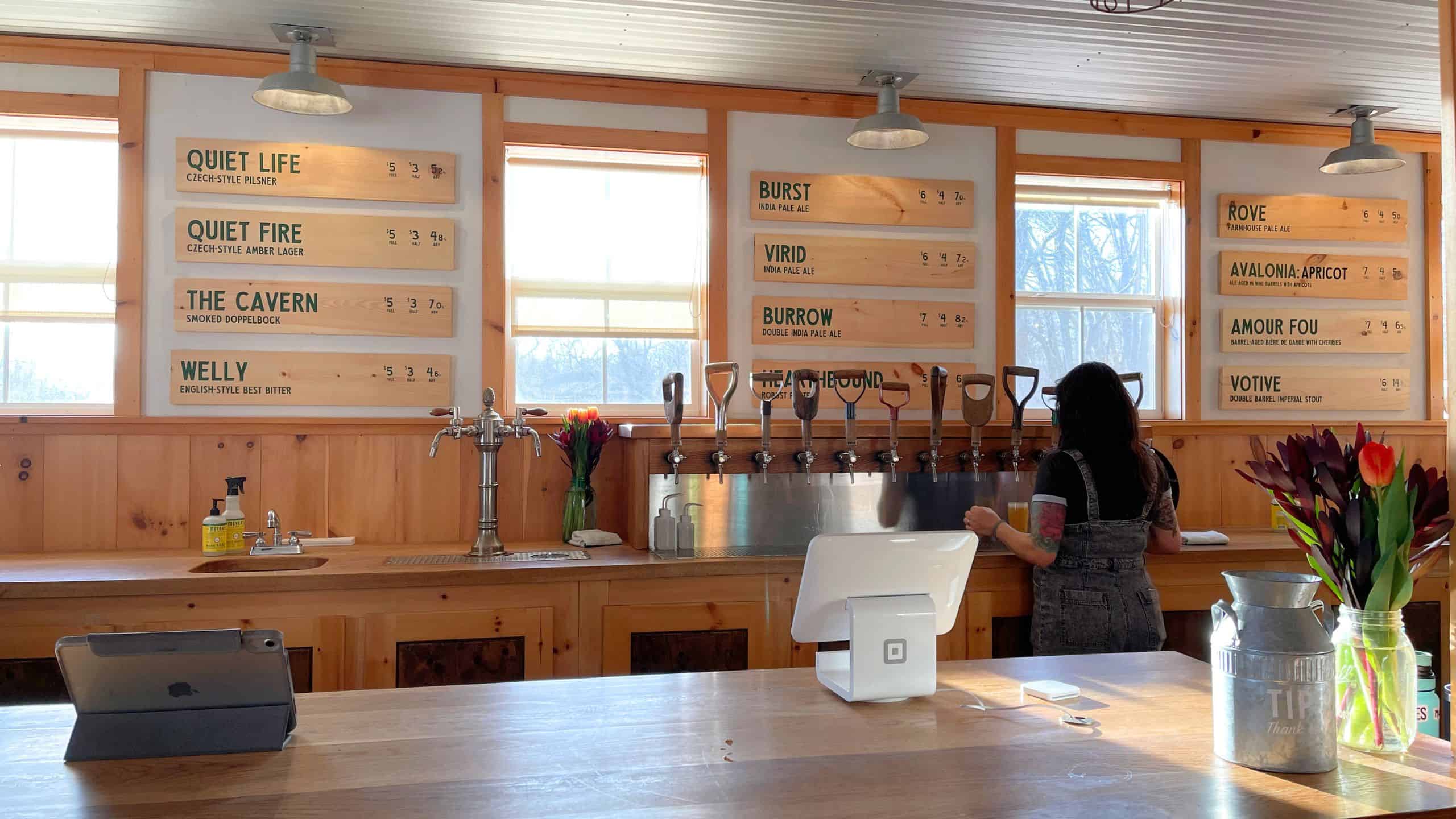 A woman stands behind a wooden bar with multiple beer taps; beer names and prices are listed on wall-mounted boards above her.