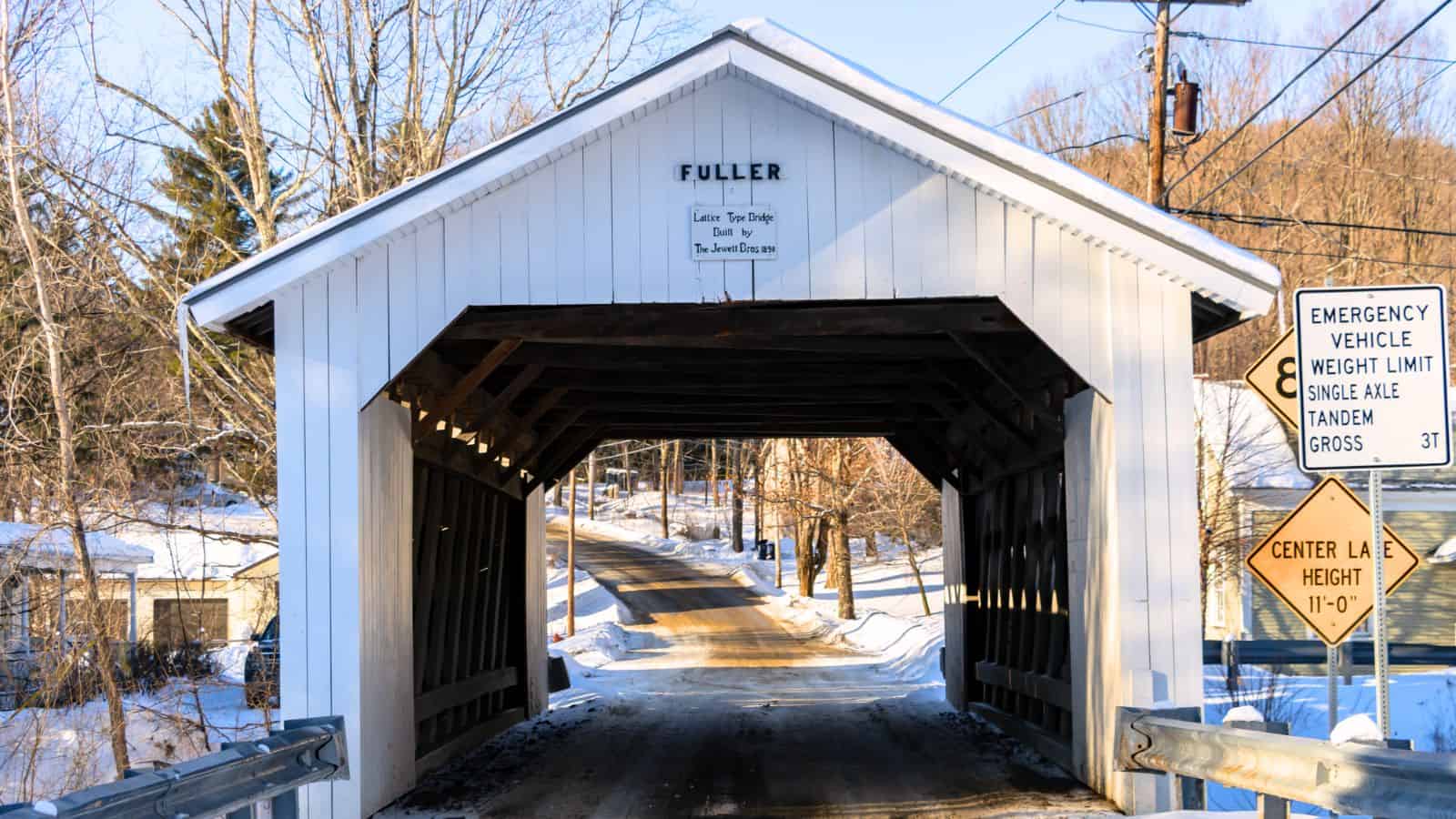A white wooden covered bridge named "Fuller," one of the classic New England covered bridges, spans a snow-lined rural road, with traffic and weight limit signs posted nearby.