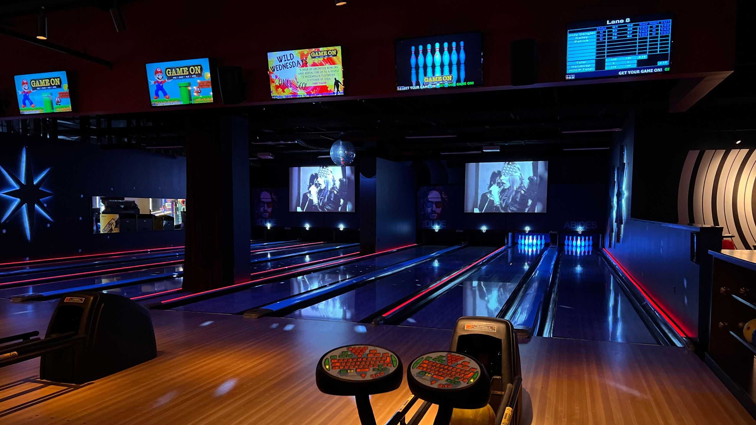 A bowling alley with several lanes, glowing red lights, overhead score screens, and large black-and-white movie projections on the back wall.