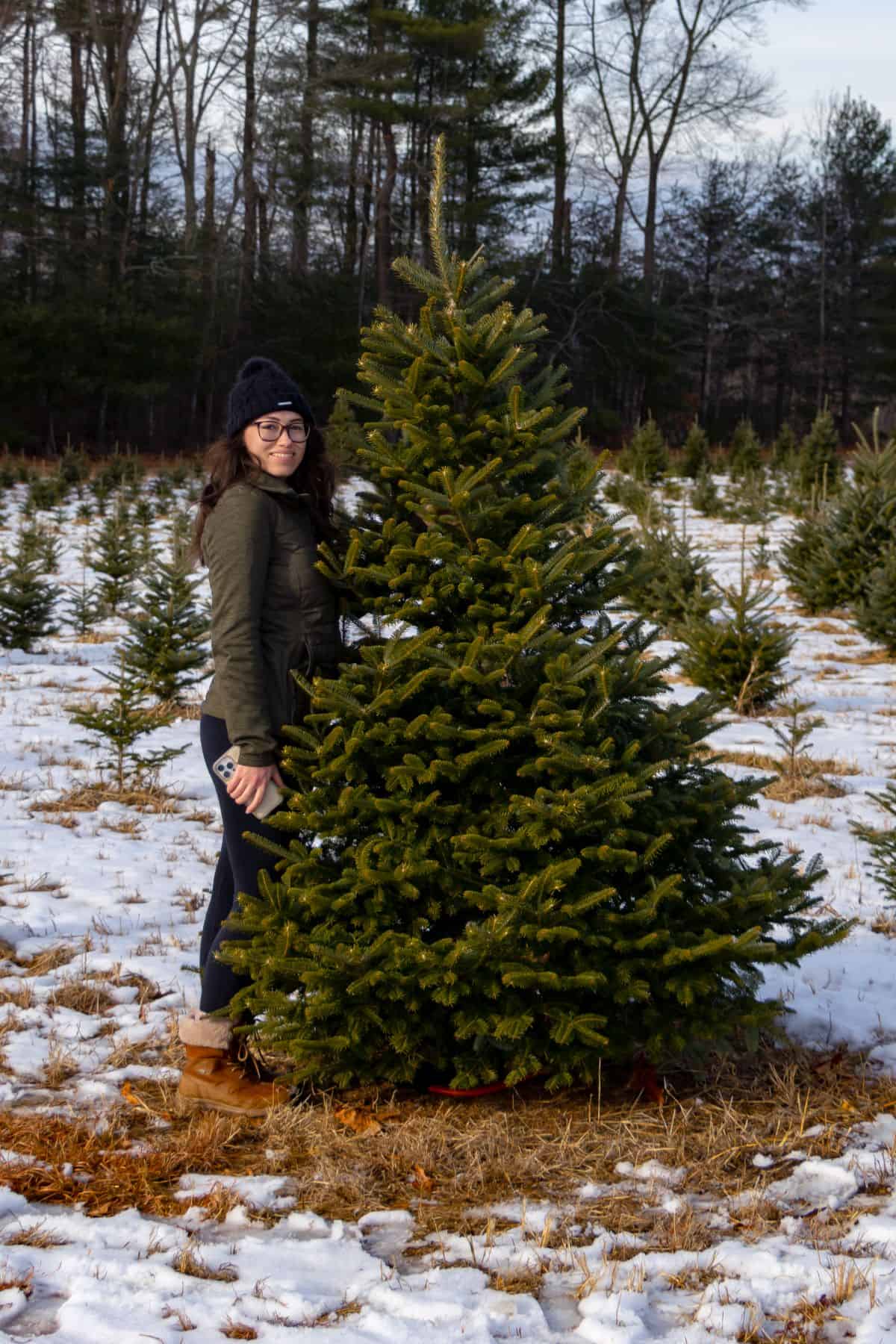 A person in winter clothing stands next to a pine tree at Geer Tree Farm's snowy grounds, holding a phone and looking at the camera.