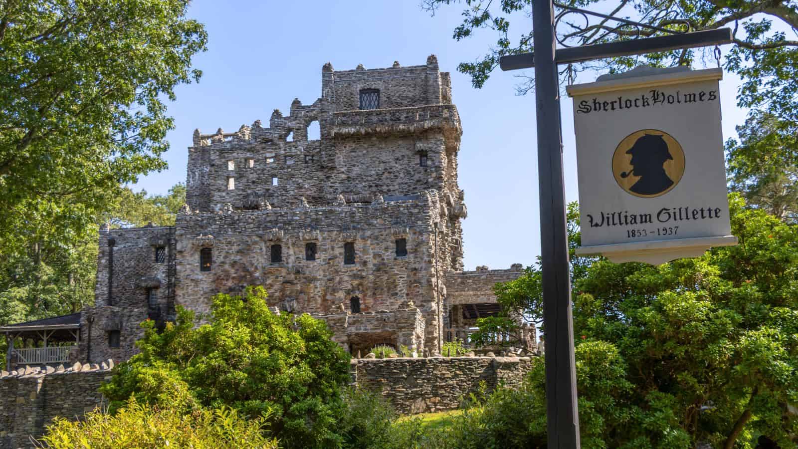 Stone castle with rugged walls surrounded by greenery; sign in foreground reads "Sherlock Holmes, William Gillette, 1853-1937" with a silhouette illustration.