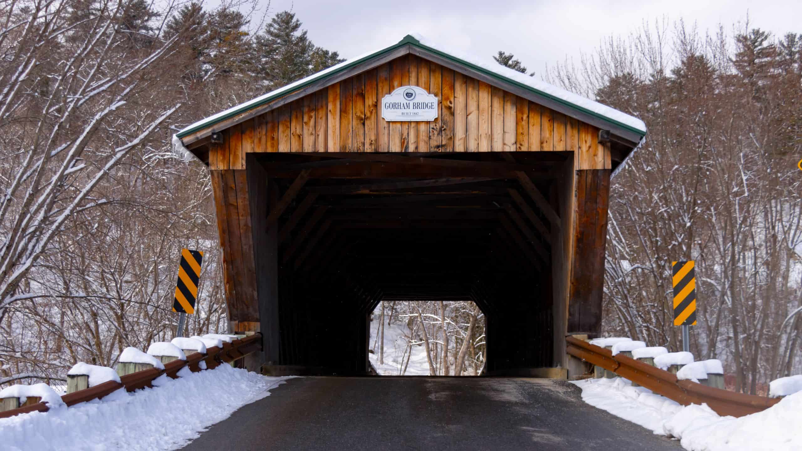 A wooden covered bridge labeled "Gorham Bridge" spans a snow-covered road, with trees and guardrails on either side.